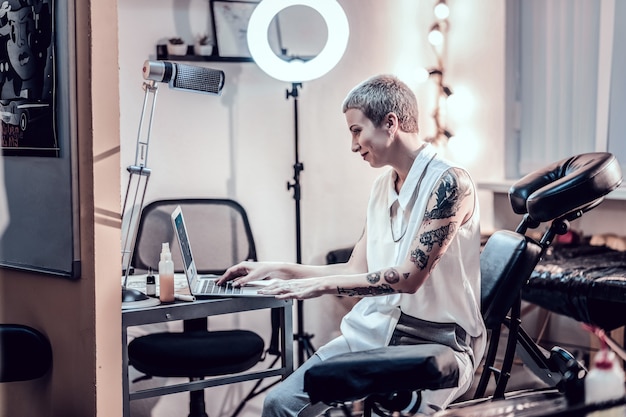 Artista concentrado. Mulher sorridente com corte de cabelo de menino trabalhando com seu laptop enquanto está sentada em uma poltrona especial