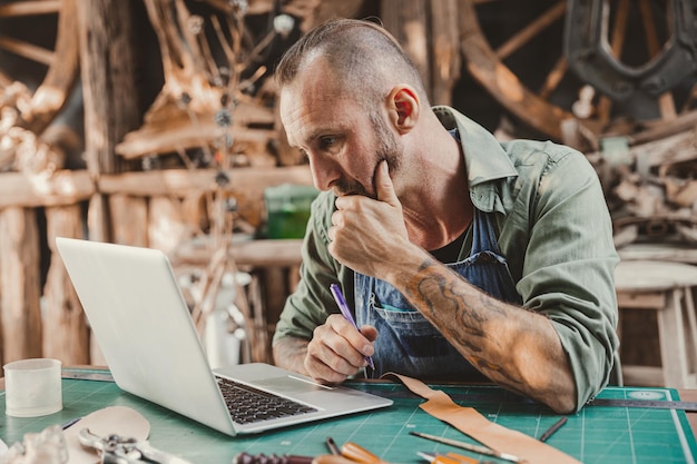 Artista de cuero hipster mirando la pantalla de la computadora portátil pensando seriamente en aprender o contactar al cliente