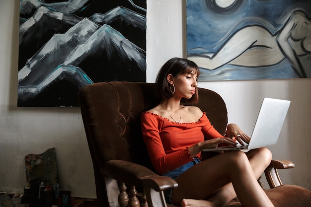 Artista de bela jovem sentada na cadeira do estúdio de arte, relaxando, usando o laptop