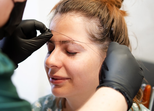 Artista de maquiagem arrancando as sobrancelhas com uma linha Tratamento profissional para o rosto.