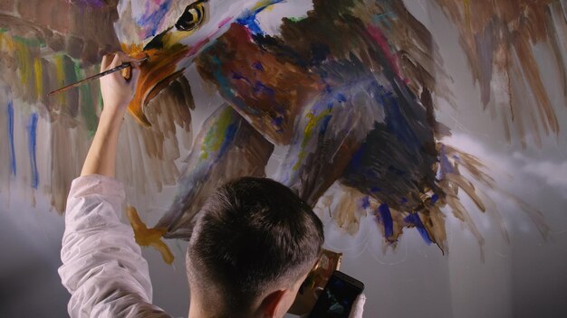 Artista desenhista desenha uma águia na parede Artesão decorador pinta imagem com cor de óleo acrílico olhando para esboço no telefone Magia negra cinematográfica