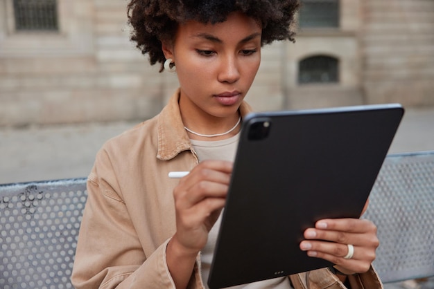 Artista digital utiliza tableta de pantalla gráfica trabaja de forma remota en un lugar público utiliza un lápiz óptico vestido con poses de chaqueta casual en un banco contra el fondo borroso al aire libre. Diseñadora creativa con gadget