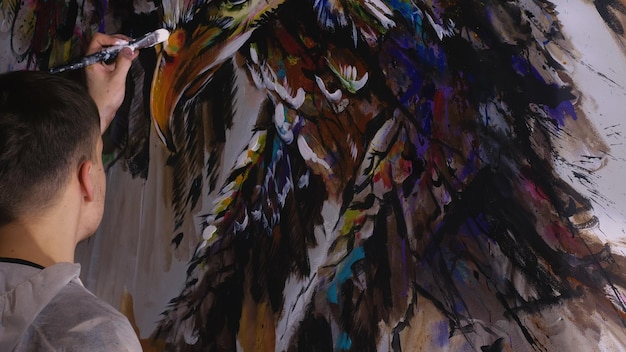 Artista diseñador dibuja un águila en la pared Artesano decorador pinta un cuadro con acrílico de color al óleo Pintor vestido con una capa de pintura Aspecto cinemático de magia oscura interior