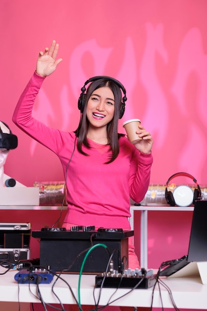 Artista feliz tocando som techno no console do mixer profissional enquanto bebe café, curtindo tocar músicas à noite no clube. Músico asiático criando performance musical com música remix