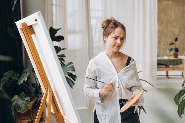 Artista femenina atractiva sosteniendo el pincel y la paleta de colores en las manos y mirando a un lado