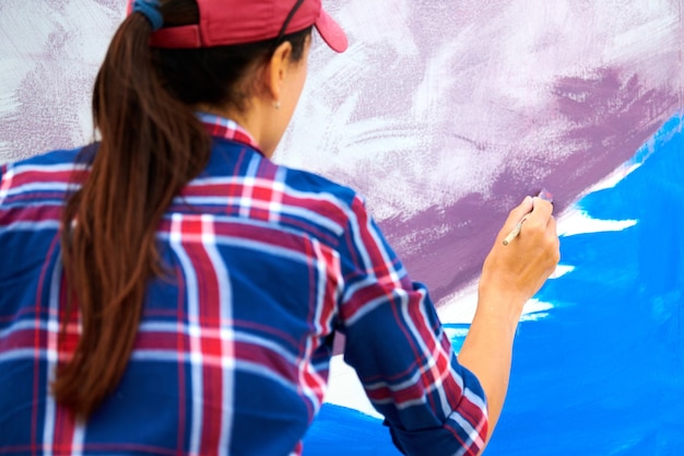 Foto artista femenina pintando sobre lienzo de madera en el festival al aire libre, joven pintora adulta creando una imagen abstracta en colores morados, violetas, azules y blancos, pincel en movimiento, vista posterior
