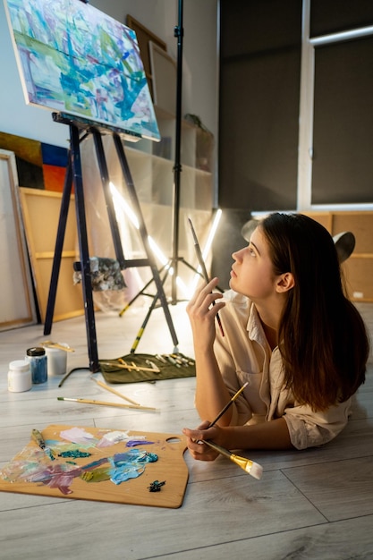 Artista femenina Proceso creativo Escuela de bellas artes Encuentra inspiración Linda mujer tendida en el piso con colores de pintura de paleta y pinceles que buscan obras de arte abstractas en el interior del estudio