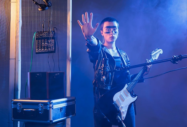 Artista femenina que muestra la señal de alto con la palma, haciendo un símbolo de ruptura serio en el estudio con humo y luces oscuras. Un rockero con estilo que se prepara para cantar música heavy metal con guitarra, concierto en solitario.