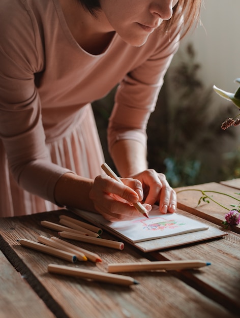 Artista feminina desenha uma composição de flores silvestres em uma panela em estilo rústico em um caderno em uma mesa com lápis.