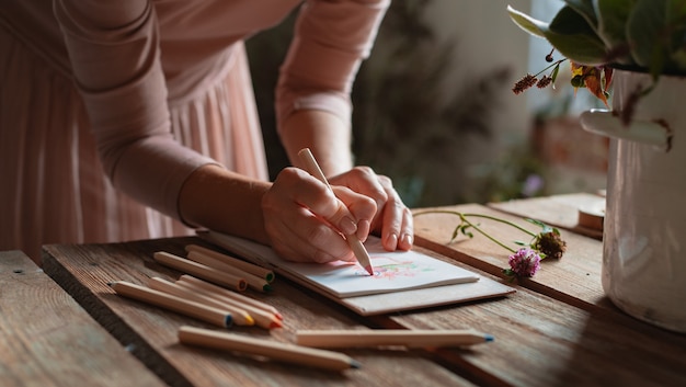 Artista feminina desenha uma composição de flores silvestres em uma panela em estilo rústico, um caderno em uma mesa com lápis.