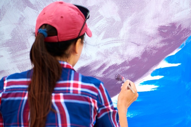 Artista feminina pintando em uma placa de tela de madeira no festival ao ar livre, uma jovem adulta pintora criando imagens abstratas nas cores roxa, violeta, azul e branca, movendo o pincel, vista traseira