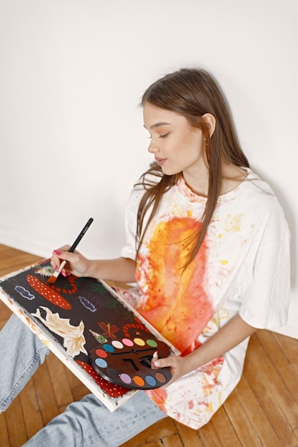 Artista feminina sentada em uma cadeira perto de parede branca segurando uma tela e pincéis