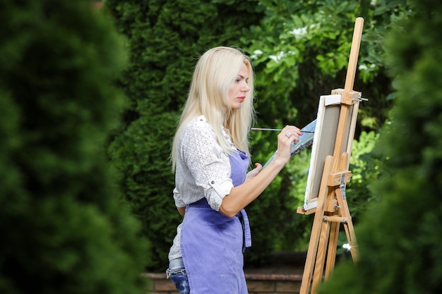 Artista hermosa mujer rubia con un pincel en la mano dibuja sobre lienzo en el jardín.