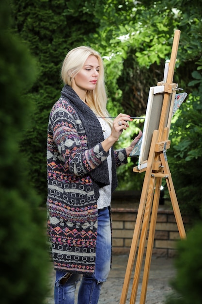 Artista hermosa mujer rubia con un pincel en la mano dibuja sobre lienzo en la naturaleza.