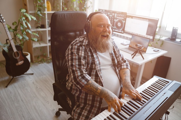 Artista hombre obeso alegre toca música en el sintetizador cerca de la mesa con monitores de computadora en estudio