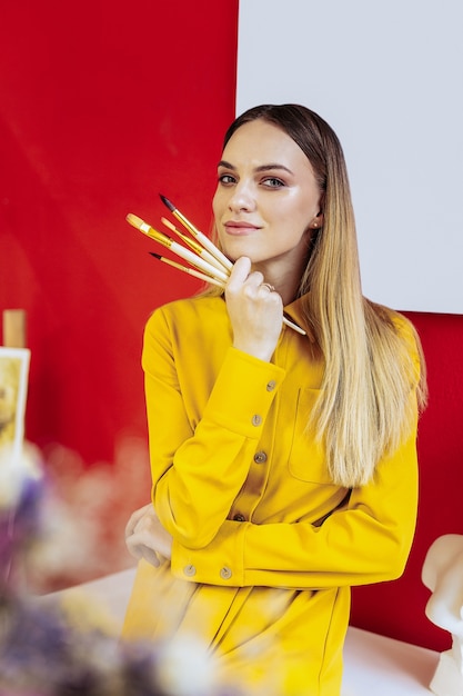 Artista inspirado. Artista atraente de olhos azuis usando um vestido amarelo e sentindo-se inspirado antes de pintar