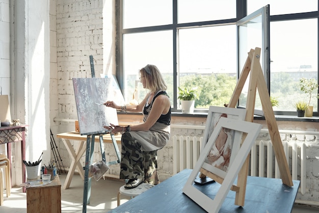 Artista loira moderna de óculos, segurando o pé no banquinho e pintando na tela usando a paleta