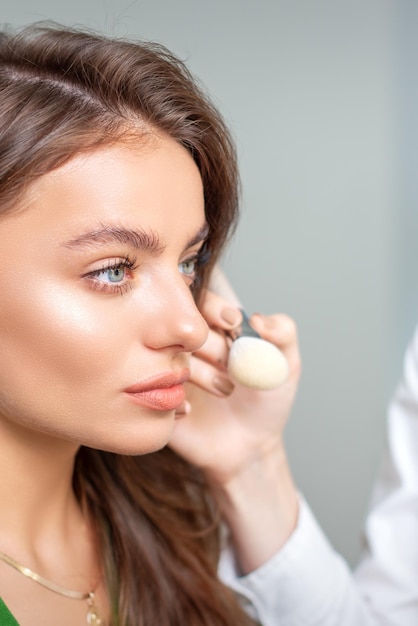 Artista de maquillaje aplicando base tonal cosmética seca en el rostro de una mujer joven con la herramienta de pincel en un salón de belleza