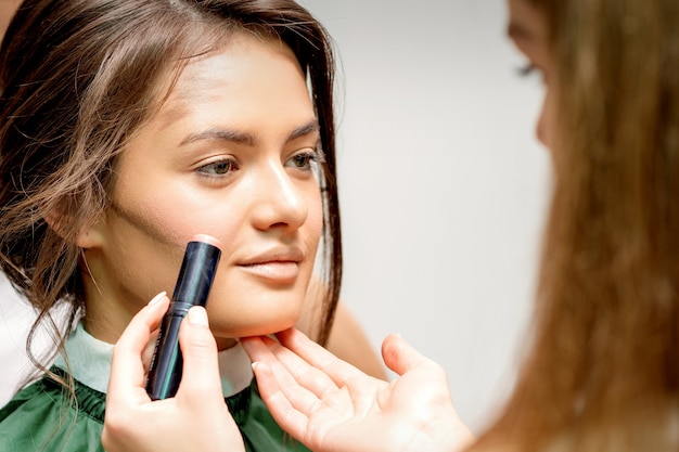 Artista de maquillaje aplicando crema blush stick foundation en la mejilla de la joven mujer caucásica