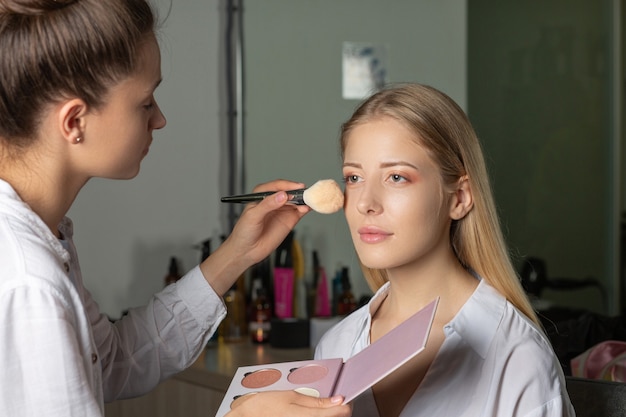 Artista de maquillaje aplicando resaltador a un cliente en el salón de belleza. Mujer trabaja con paleta de maquillaje y pincel natural