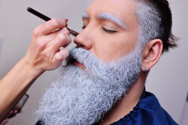 El artista de maquillaje maquilla a un hombre con barba blanca o gris.