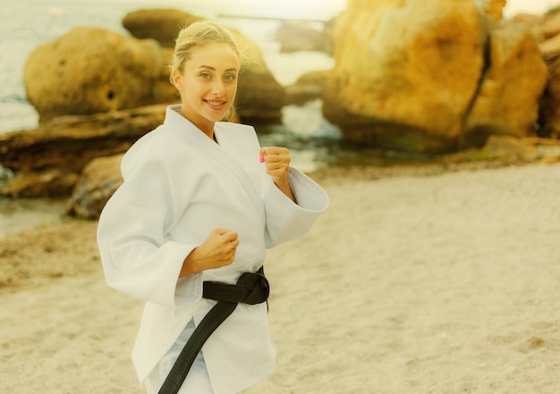 Artista marcial femenina atractiva en un kimono blanco con un cinturón negro se encuentra en posición de combate en la playa salvaje