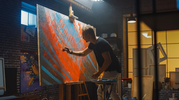 Artista masculino talentoso dibuja enérgicamente y violentamente con pincel crea una obra maestra moderna del estudio creativo de pintura al óleo con un gran lienzo de colores llamativos