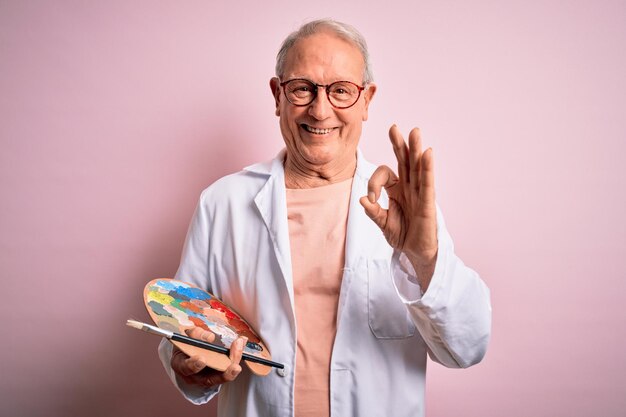 Foto un artista mayor de pelo gris pintando con paleta de pintor sobre fondo rosa haciendo el signo correcto con los dedos símbolo excelente