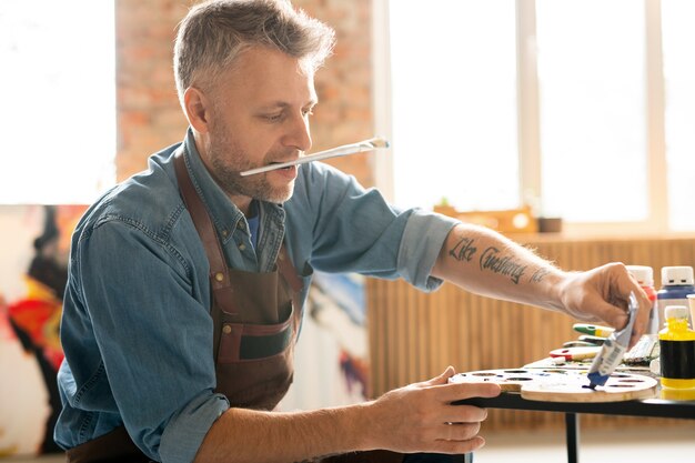 Artista de mediana edad con pincel en la boca sentado junto a la mesa en el taller y presionando pintura de aceite azul del tubo en la paleta