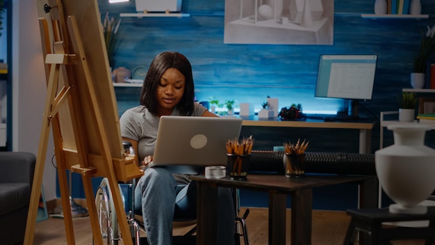 Artista negro con discapacidad escribiendo en una computadora portátil que busca inspiración en la sala de arte en casa. Mujer creativa de etnia afroamericana en silla de ruedas usando dispositivo y tecnología