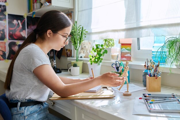 Artista de niña creativa adolescente dibujando con un lápiz sentado en la mesa en casa usando maniquí de madera Creatividad escuela hobby ocio estilo de vida adolescencia juventud concepto