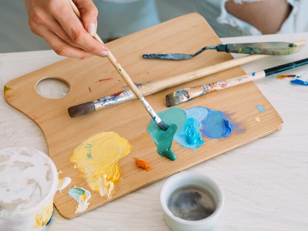 Artista no local de trabalho Pintar arte Talento de habilidade Pintor profissional mãos de mulher misturando tinta acrílica de cor azul amarela com pincel na paleta de madeira com ferramentas