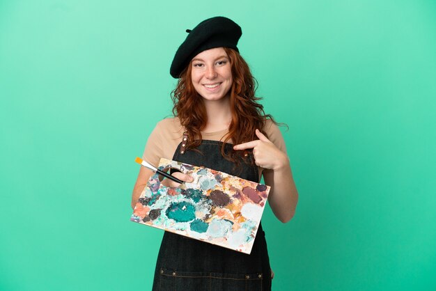 Artista pelirroja adolescente sosteniendo una paleta aislada sobre fondo verde con expresión facial sorpresa