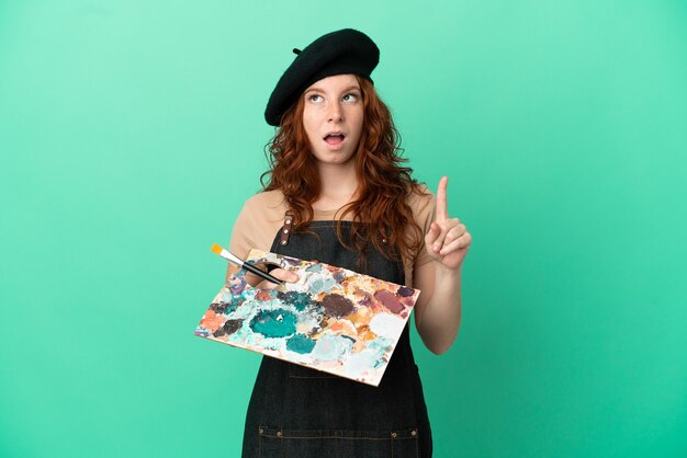 Artista pelirroja adolescente sosteniendo una paleta aislada sobre fondo verde con la intención de darse cuenta de la solución mientras levanta un dedo