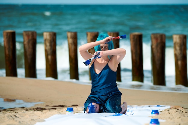 Artista de performance mujer de pelo azul artístico en vestido azul oscuro manchado con pintura gouache azul mientras está sentado en la playa. Pintura corporal y concepto de arte corporal, arte de actuación expresiva al aire libre