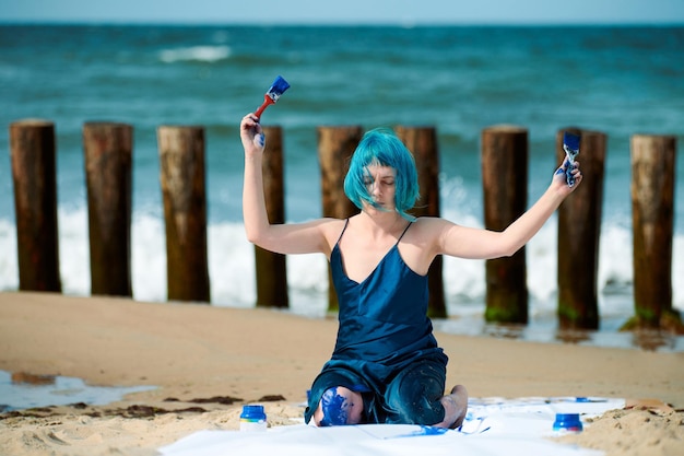 Artista de performance mujer de pelo azul artístico en vestido azul oscuro manchado con pintura gouache azul mientras está sentado en la playa. Pintura corporal y concepto de arte corporal, arte de actuación expresiva al aire libre