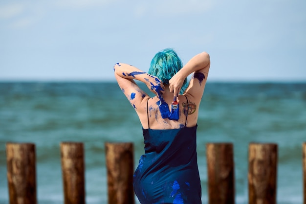 Artista performática de mulher de cabelo azul, em um vestido azul escuro manchado com pintura a guache índigo em seu corpo com pincéis na praia, vista traseira. pintura corporal criativa, conceito de arte corporal, ao ar livre