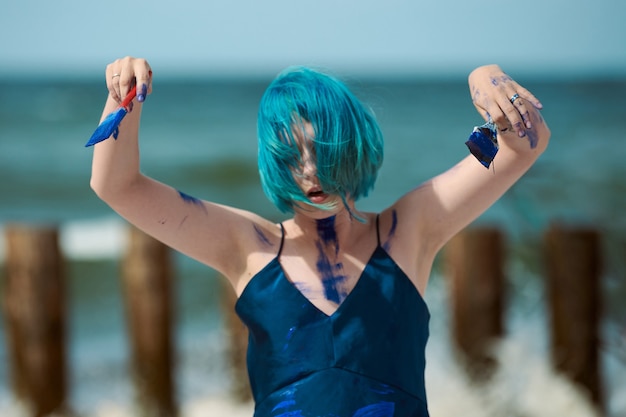 Artista performática de mulher de cabelo azul, num vestido azul escuro manchado com tintas de guache azuis, pintando e dançando na praia. Conceito de pintura corporal e arte corporal, arte performática expressiva ao ar livre