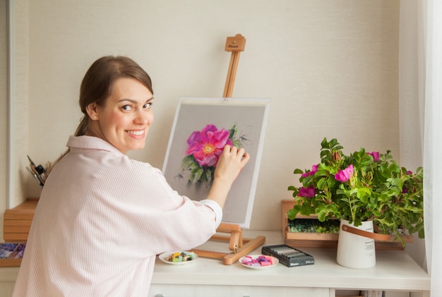 Artista positivo joven hermosa chica sonriente trabaja en estudio selecciona colores de cama para trabajar con flor rosa mosqueta sentado en la mesa con un caballete y un ramo. La creatividad conceptual como profesión