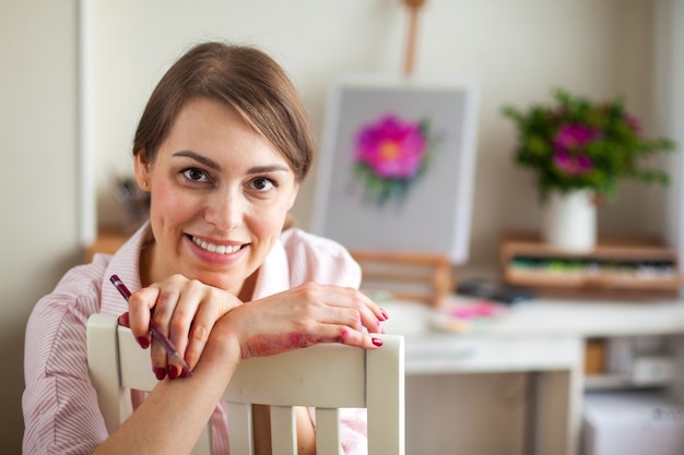 Artista positivo joven hermosa niña sonriente trabaja en estudio selecciona colores de cama para trabajar con flor rosa mosqueta sentado en la mesa con un caballete y un ramo. La creatividad conceptual como profesión