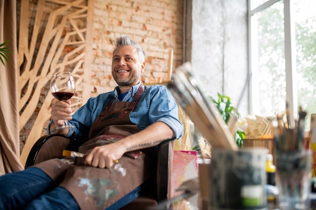 Artista profesional feliz con copa de vino brindando por el éxito después de terminar la nueva obra maestra en el estudio