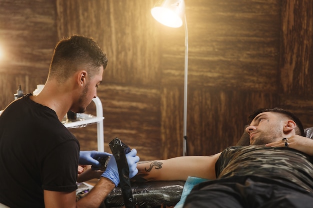 Artista tatuador profesional haciendo tatuaje en el brazo de un joven por máquina con tinta negra