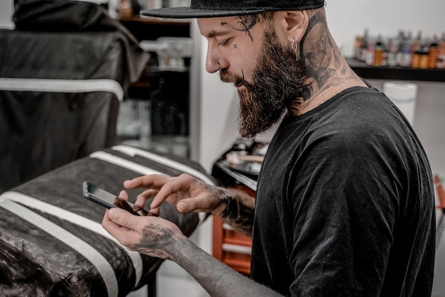 Artista del tatuaje masculino joven con barba comprobando su teléfono sentado en el sofá en el lugar del taller.