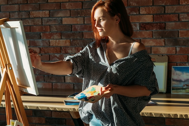 Artista en el trabajo. Arte de estudio. Mujer pelirroja pensativa con paleta de madera dibujo sobre lienzo.