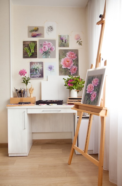 Artistas casa lugar de trabajo mesa de madera clara para pintar con caballete y hermosas obras de arte con flores colgadas en la pared y herramientas de pintura. Concepto de creatividad y afición