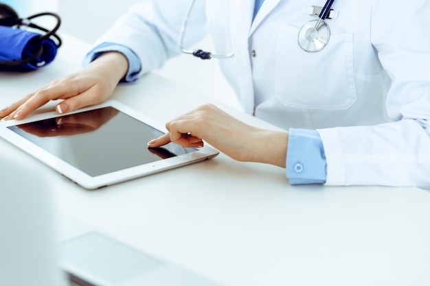 Arzt Arbeitstisch. Ärztin, die Tablet-Computer verwendet, während sie in der Nähe des Krankenhausbüros sitzt. Gesundheits-, Versicherungs- und Medizinkonzept.