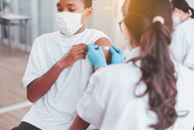 Arzt, der einen afrikanischen jungen in die gesichtsmaske mit klebeverband legt, nachdem er während einer coronavirus- oder covid-19-pandemie im krankenhaus geimpft wurde, impfkonzept