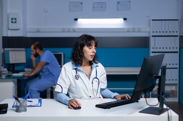 Arzt, der im Büro auf der Computertastatur tippt und bis spät in die Nacht arbeitet. Ärztin mit Stethoskop mit Blick auf die Monitoranzeige für das Gesundheitssystem und die Unterstützung nach Stunden