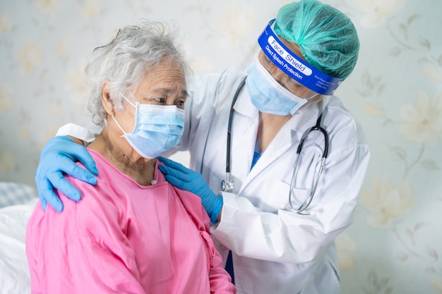 Arzt, der Stethoskop verwendet, um asiatische ältere Patientin zu überprüfen, die eine Gesichtsmaske trägt, um Covid-19 Coronavirus zu schützen.