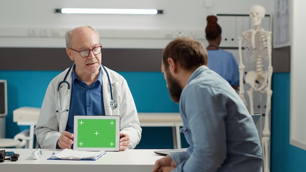 Arzt hält digitales Tablet mit horizontalem Greenscreen bei Termin mit männlichem Patienten in Arztpraxis. Isolierter Chroma-Key-Hintergrund mit leerer Mockup-Vorlage und Copyspace.